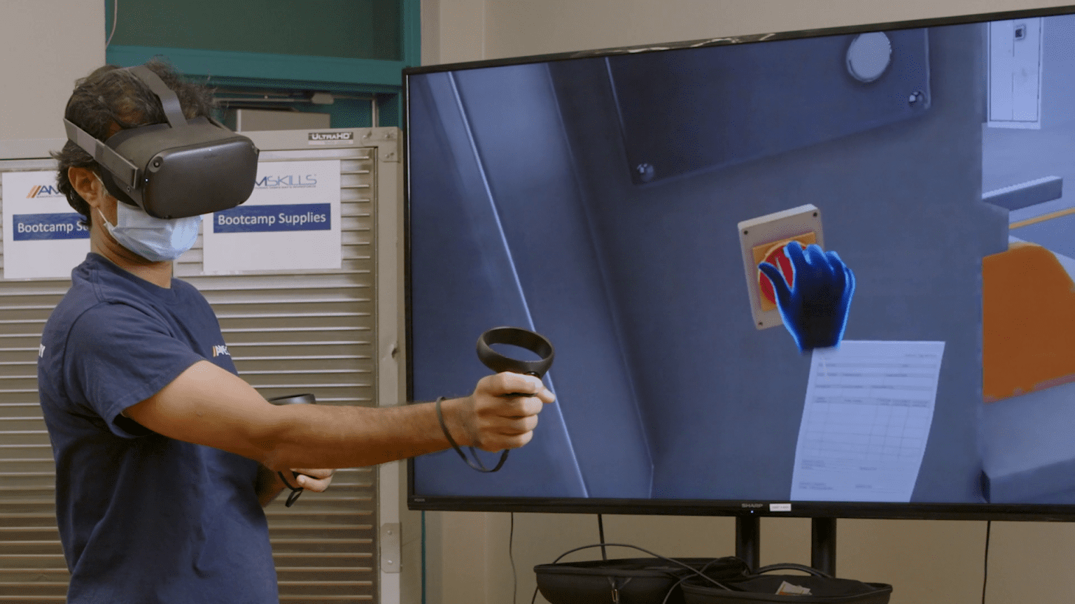 A student in a blue shirt using a VR headset and hand controls; a monitor screen in the same shot shows a VR hand in a simulation.