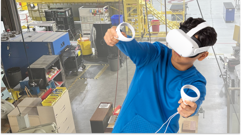 A person in a blue shirt with a headset on, using hand controllers, with a virtual reality background showing a warehouse floor.